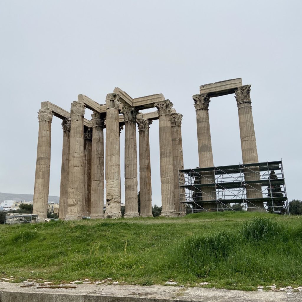 a group of pillars on a grassy hill with Temple of Olympian Zeus, Athens in the background