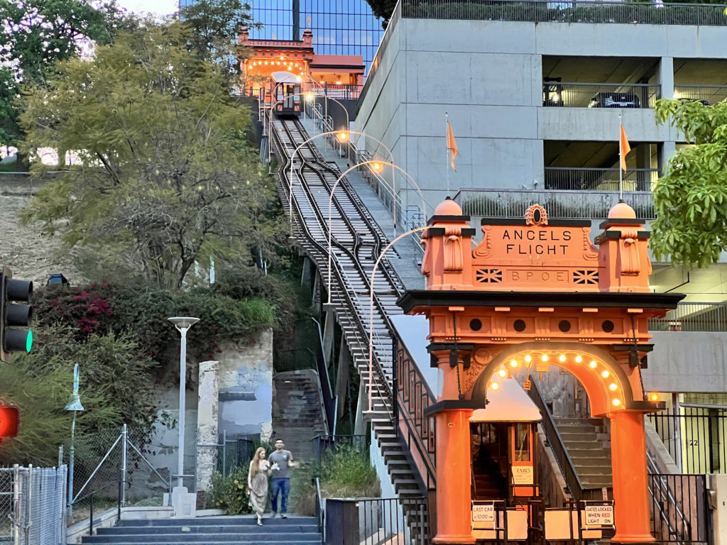 an orange archway with lights and a couple of people walking down a hill with Angels Flight in the background